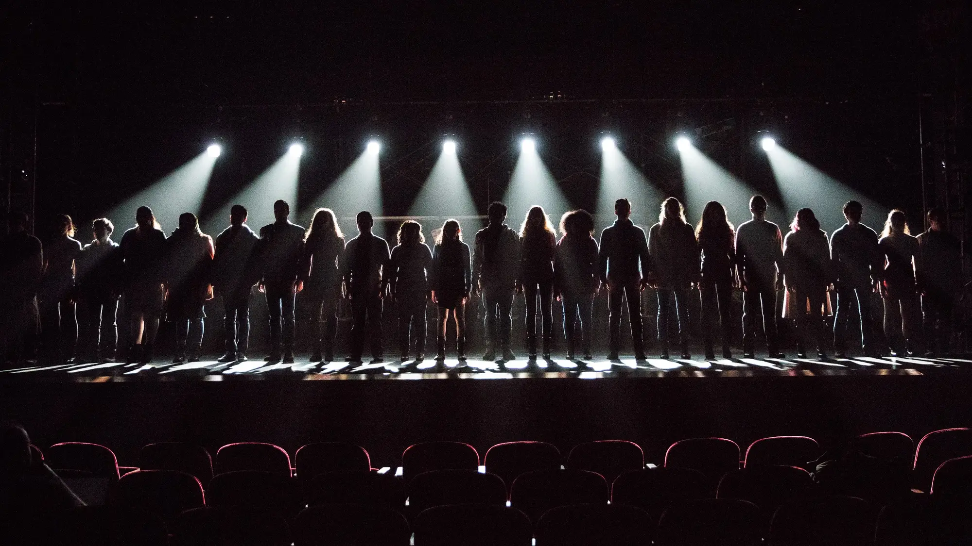 A line of actors stand on stage backlit by stage lights. 