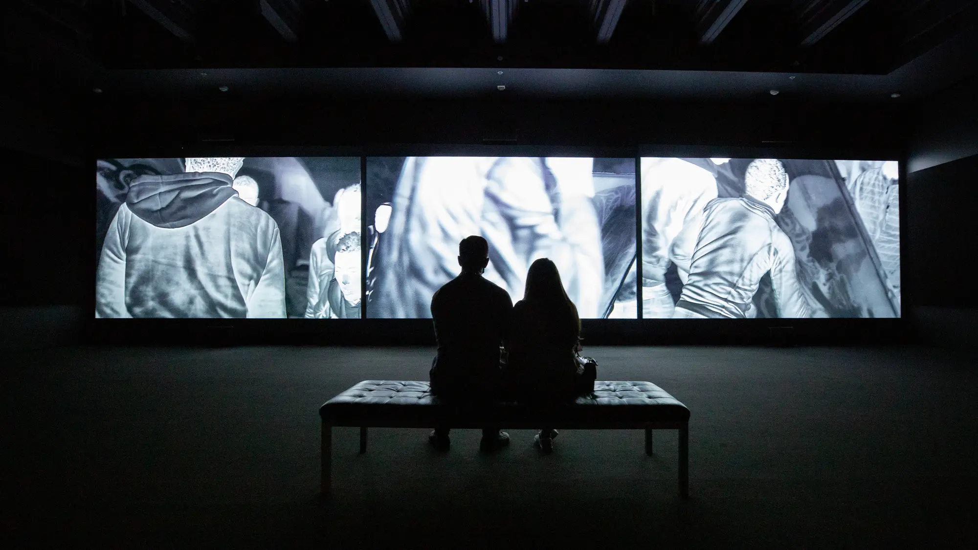 A man and woman sit on a bench looking at floor-to-ceiling illuminated photographs. 