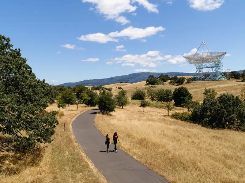 Two people walking along the paved path surrounded by trees and the Stanford Dish.