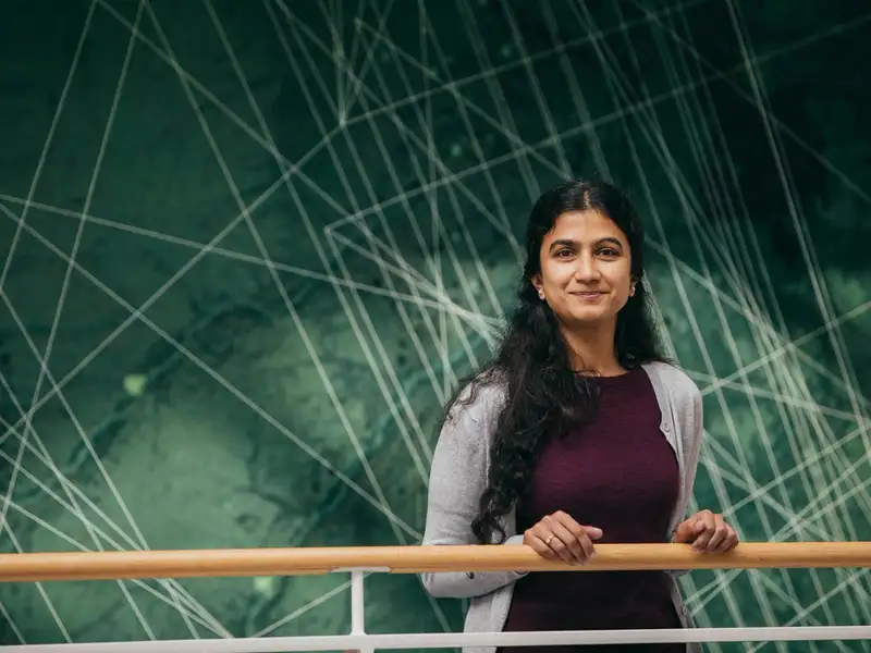 Aditi Sheshadri stands at a railing, smiling in front of a large green mural with intersecting white lines.