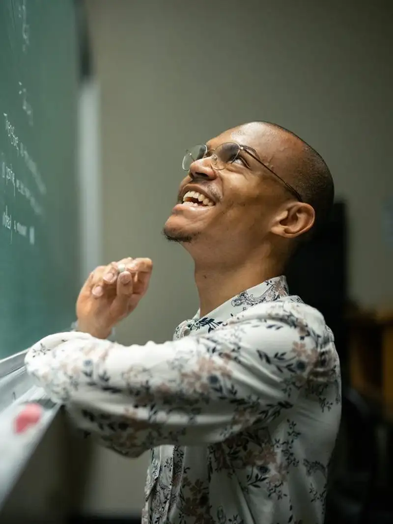 Jadon Geathers smiles and looks up at a chalkboard, holding chalk mid-thought while working through a problem in a classroom.