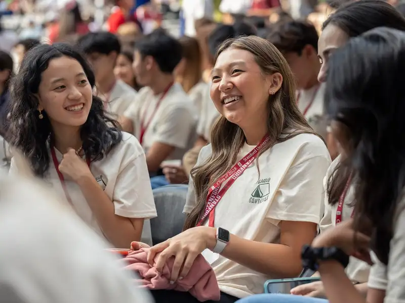 Smiling Stanford students in matching cream t-shirts and red lanyards chat together while seated in a crowd at a new-student convocation event.