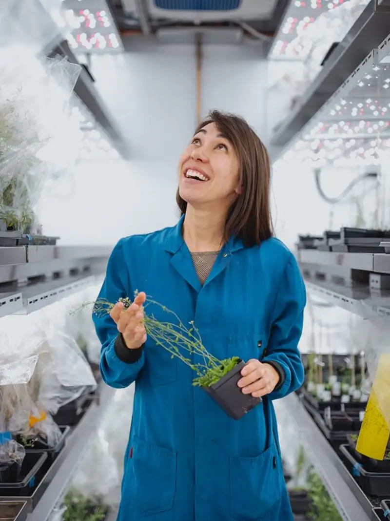 Jenn Brophy in a blue lab coat holds a small plant pot while working in a greenhouse filled with seedlings and grow lights.