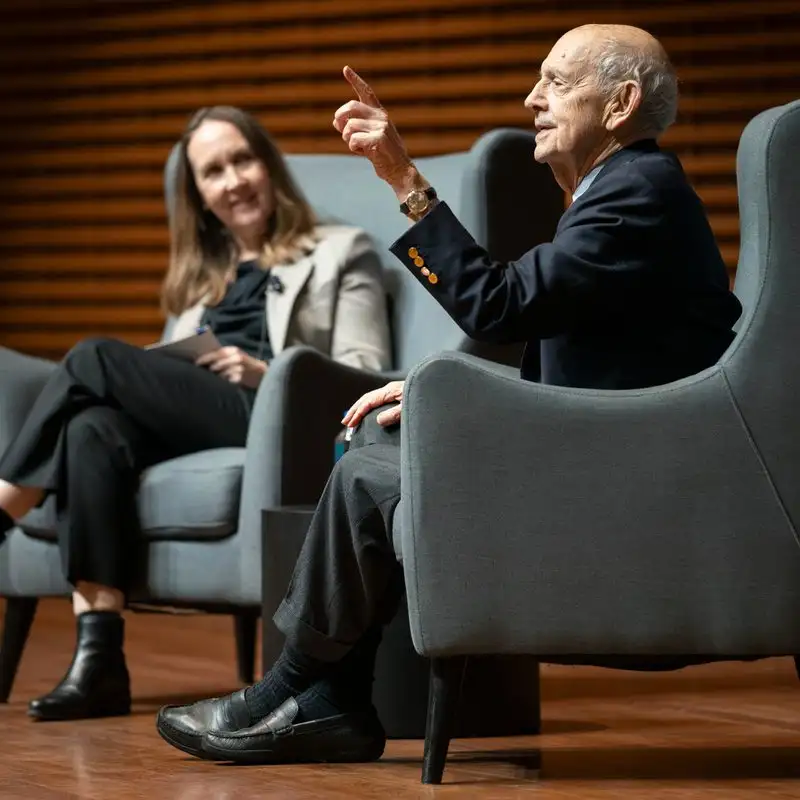 Justice Stephen Breyer gestures while speaking on stage, seated in an armchair beside Stanford University Provost Jenny Martinez, who listens during a moderated conversation.