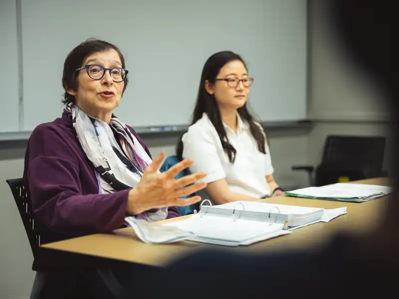 Pam Karlan gestures while speaking at a seminar table, with a student seated alongside her and open binders spread before them.