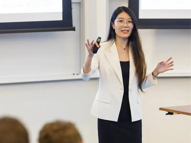 Yuyan Wang presents to an audience, holding a remote in one hand, in a classroom setting.