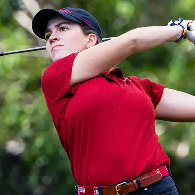Paula Martin Sampedro swings a club on the course, wearing a red polo and black pants, with greenery in the background.