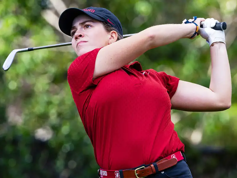 Paula Martin Sampedro swings a club on the course, wearing a red polo and black pants, with greenery in the background.