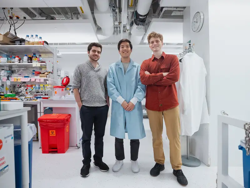 Three researchers stand together smiling in a bright, well-stocked lab, one wearing a blue lab coat.