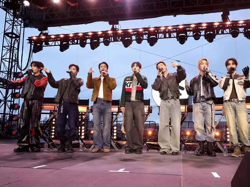 Seven performers of BTS stand in a line on an outdoor stage under bright lights during an evening concert.