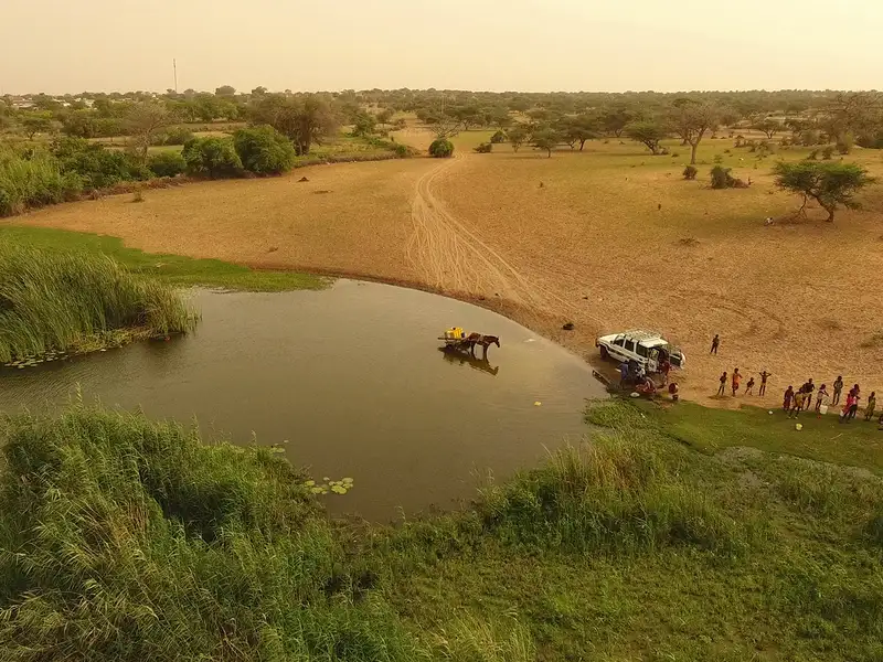 Aerial view of a riverbank with a horse and cart in shallow water, and people gathered along the shore near a parked vehicle.