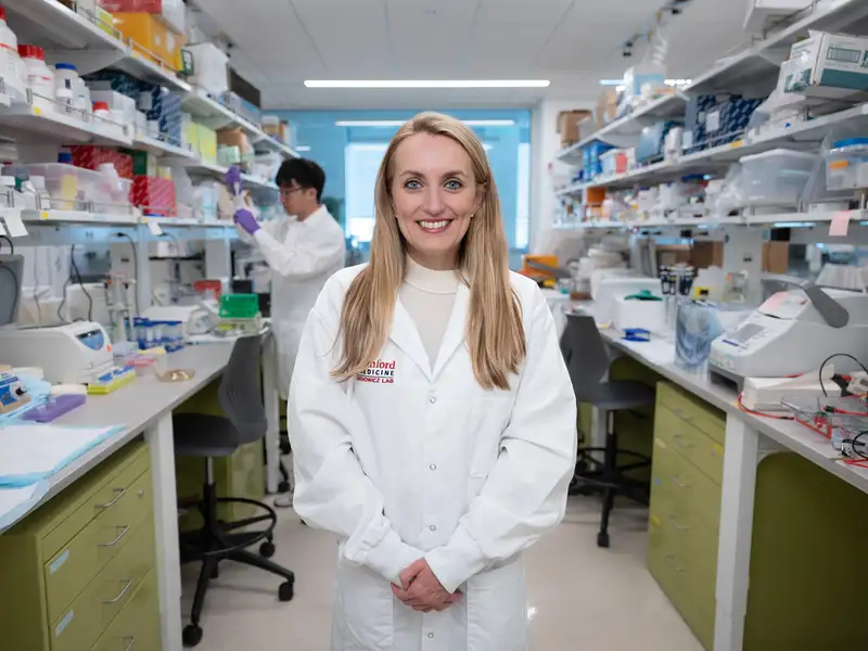 Agnieszka Czechowicz in a lab coat standing in a laboratory filled with equipment and materials, while a colleague works in the background.
