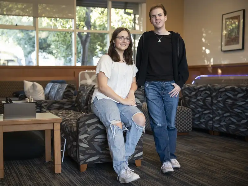 Stella Vangelis and Peter Bennett pose together in a casual campus lounge, one seated and one standing, with sunlit trees visible through the windows behind them.