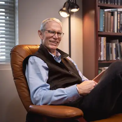 Josh Ober sits relaxed in a leather armchair, smiling, with a warmly lit bookshelf filling the background.