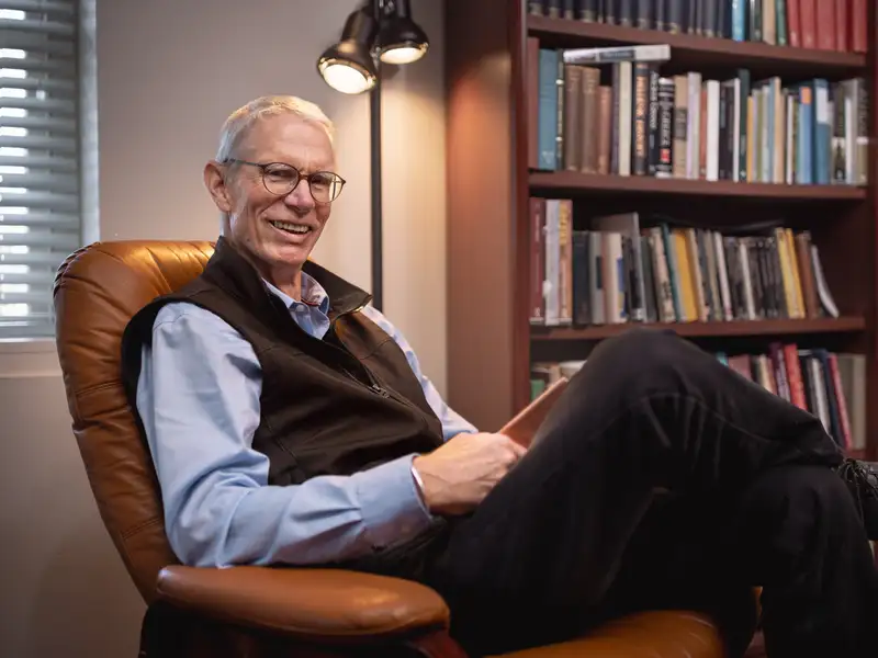Josh Ober sits relaxed in a leather armchair, smiling, with a warmly lit bookshelf filling the background.