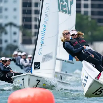 Teams of sailors compete in a race, leaning into their boats to catch the wind, with a city skyline in the background.
