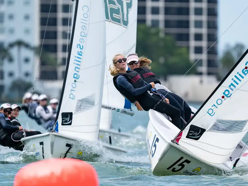 Teams of sailors compete in a race, leaning into their boats to catch the wind, with a city skyline in the background.