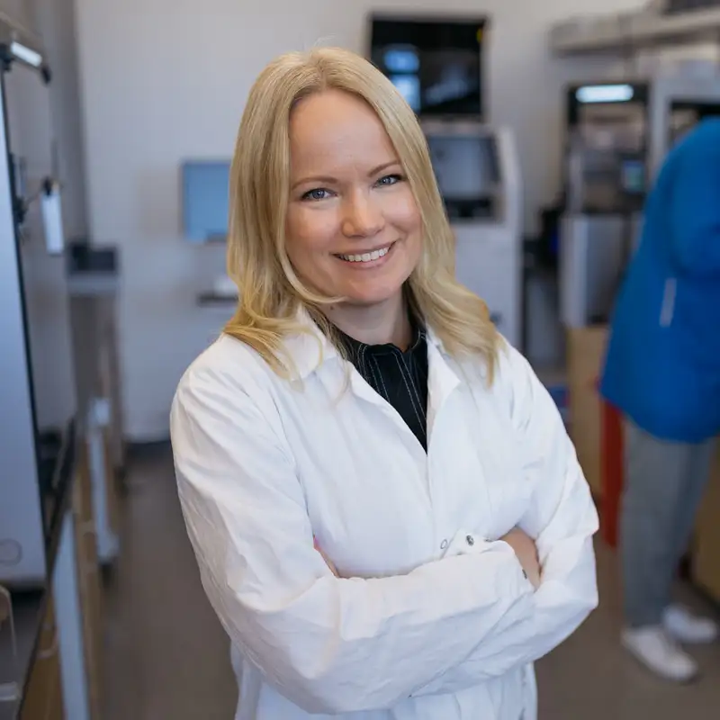 Emma Lundberg in a lab coat standing and posing for a photo, while a colleague examines equipment in the background.