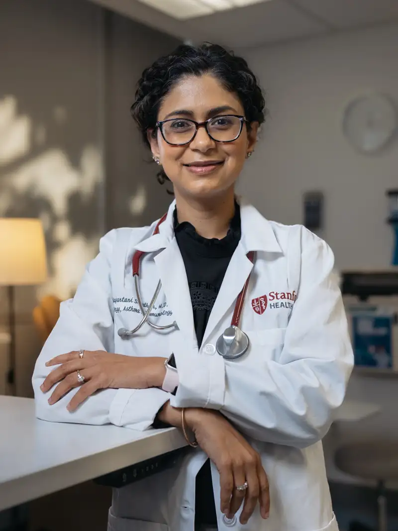 Tina Sindher in a lab coat stands in a clinic, showcasing a patient care environment.