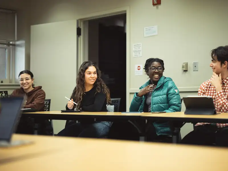 Four students sit together at a seminar table, one speaking animatedly while the others listen and smile.