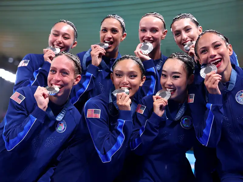 The U.S. artistic swimming team poses together, grinning and biting their silver medals at the Paris 2024 Olympics.