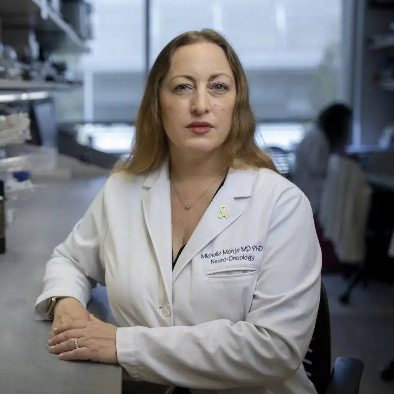 A woman wearing a white lab coat with "Michelle Monje MD PHD" on it sits at a lab bench and looks at the camera