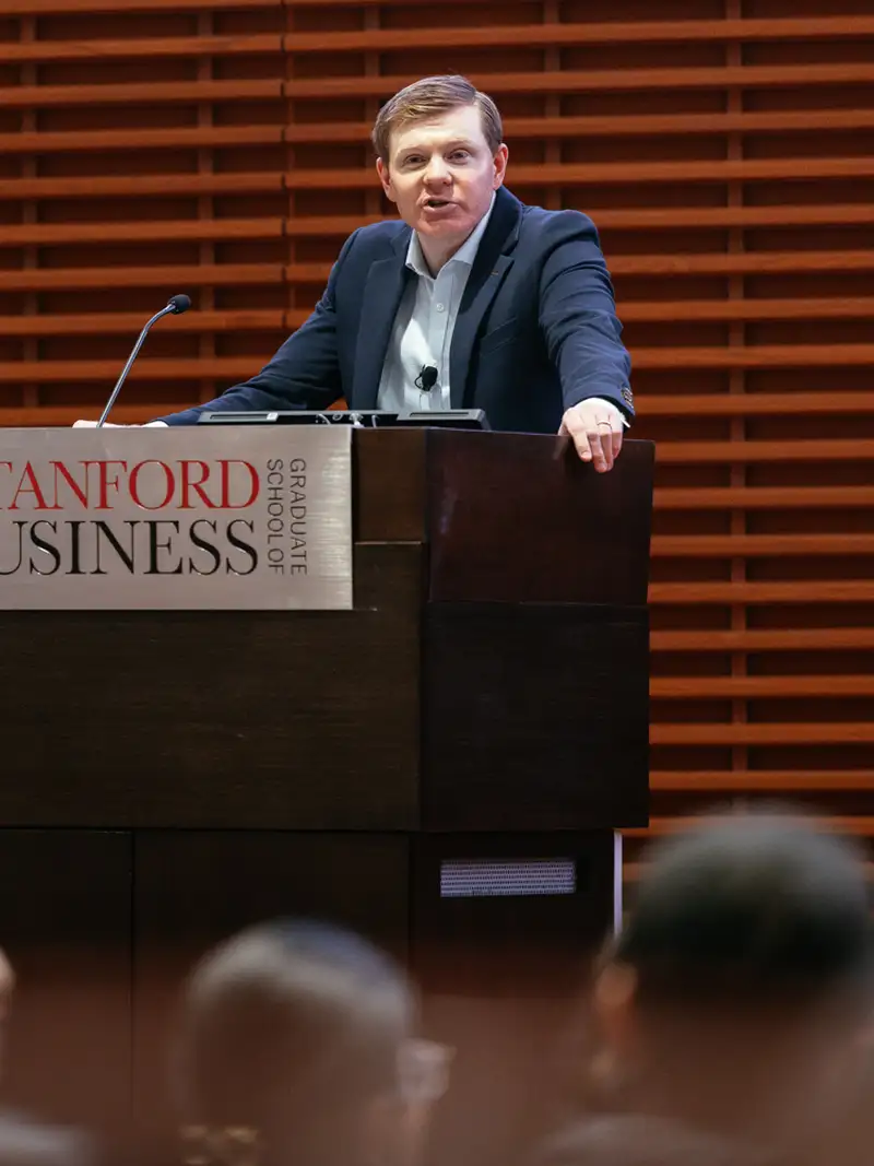 An adult male stands at a podium with the words "Stanford Graduate School of Business" on it and speaks to an audience