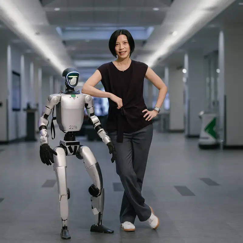 Karen Liu stands beside a humanoid robot in a modern indoor setting, both showcasing a friendly pose.