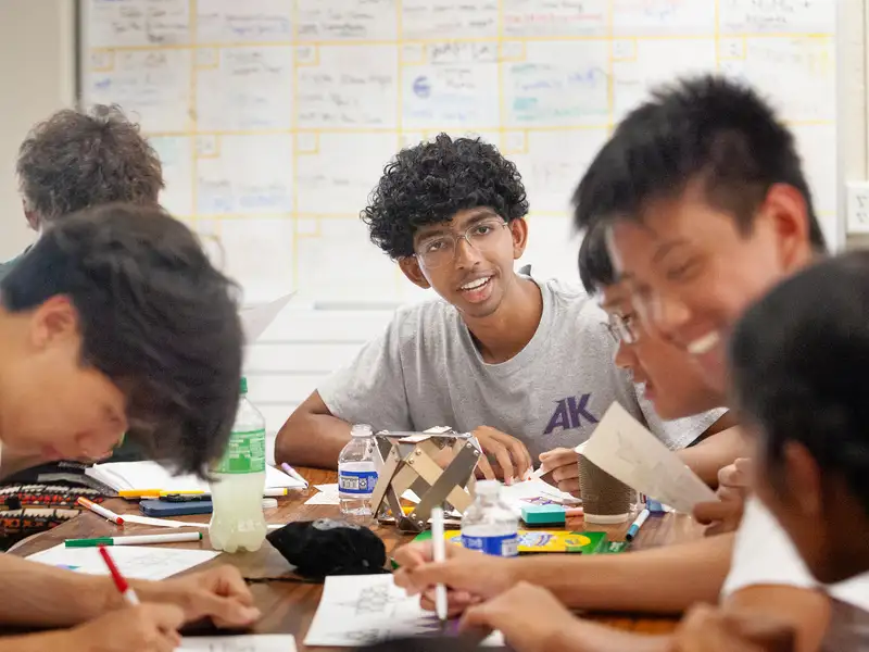 Students gather around a table, using art supplies and papers for a project while discussing ideas in an educational setting.