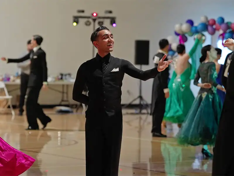 Rohit Nema in a black ballroom suit extends his arm at a dance event, among partners in colorful gowns.