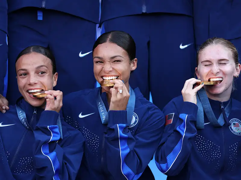 Former Stanford soccer players Tierna Davidson, Naomi Girma, and Sophia Smith celebrate winning gold by smiling and biting their gold medals.