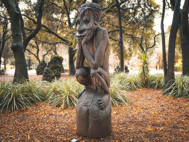 A tall carved wooden Papua New Guinea figure stands in a wooded sculpture garden among autumn leaves and grasses.