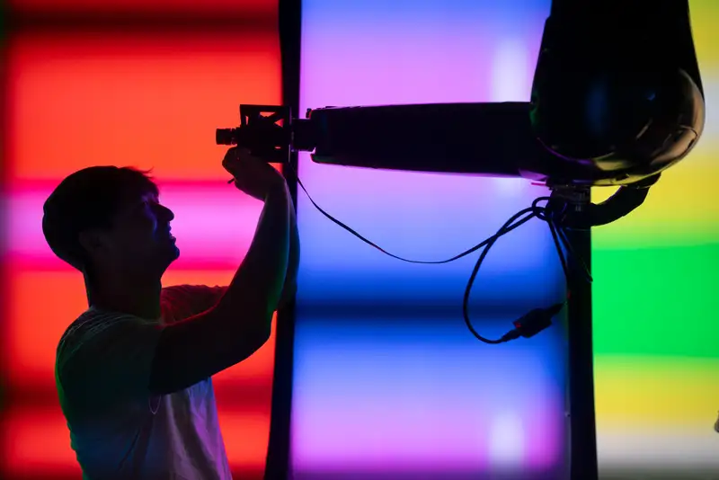 A researcher is silhouetted against vivid panels of colored light as he adjusts a sensor on a robotic arm overhead.