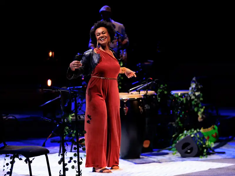 Singer Meklit, in a red jumpsuit and leather jacket, laughs while holding a microphone onstage, amid congas and trailing greenery.