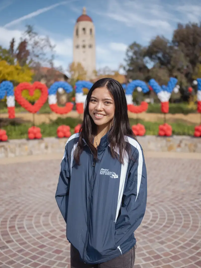 A female college student wearing a blue jacket smiles in front of balloons that spell out "Democracy Day"