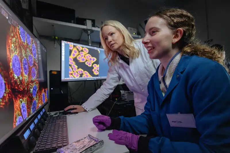 Two researchers examine vivid fluorescence microscopy images of cells displayed across two monitors in a darkened lab.