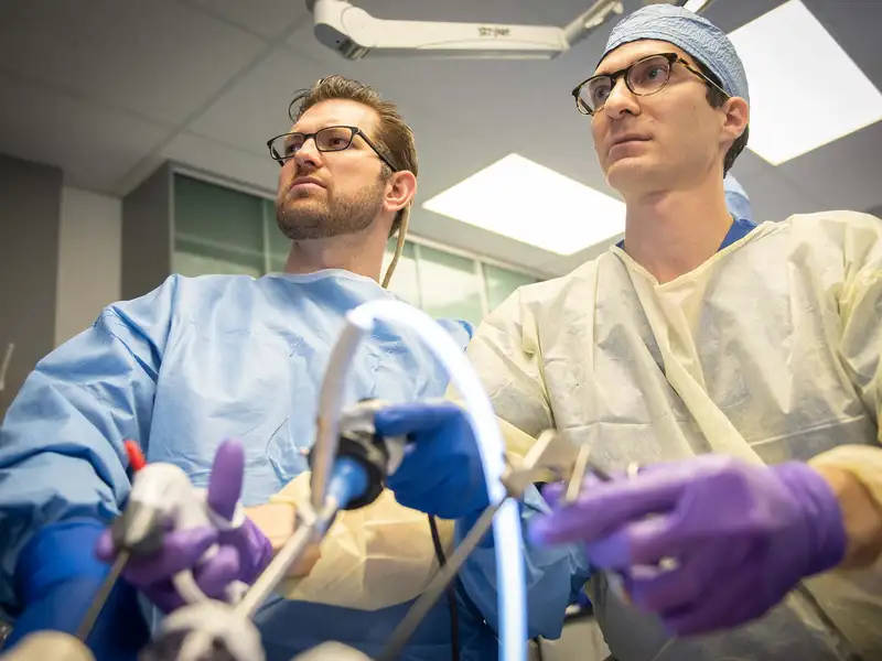 Two people in gowns and gloves look up toward a monitor while maneuvering surgical instruments, photographed from a low angle beneath the surgical tools.