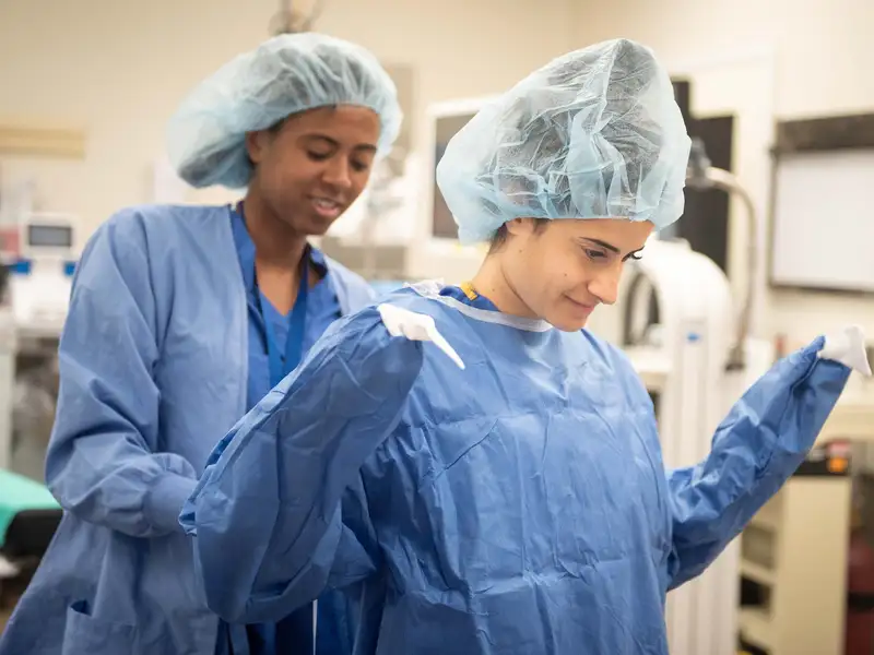 Two medical students in surgical attire assist each other in putting on protective gowns in a medical setting.