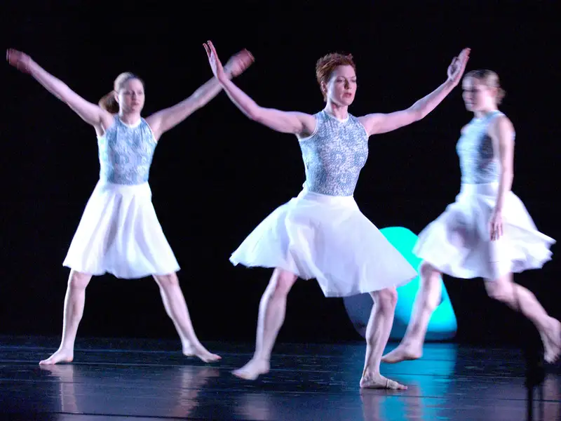 Three barefoot dancers in white skirts perform with raised arms on a dark stage, captured mid-motion with slight blur.