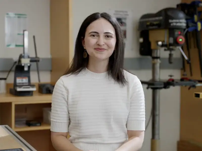 Aya Mouallem smiles at the camera in a workshop, with a drill press and clamps visible behind her.