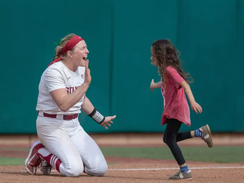 A softball player kneels with a smile and holds her hand out for a high-five as a young girl runs toward her on the field during a game.