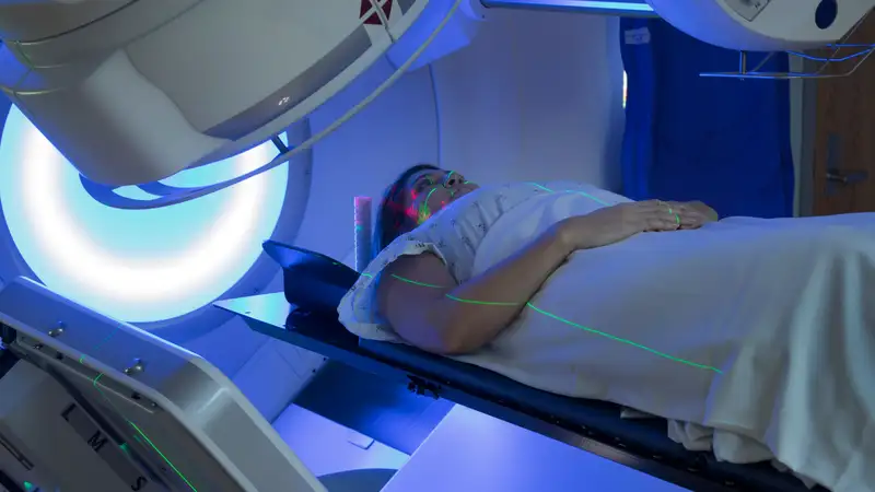 A patient wearing a hospital gown lies on her back and green laser beams go across her face and arms