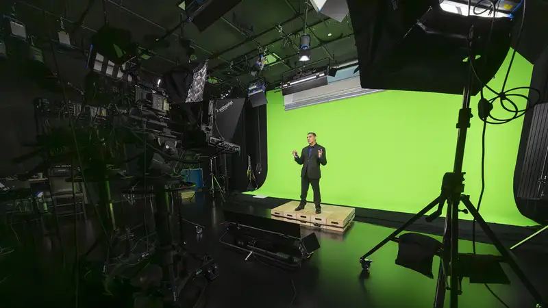 A presenter in a suit stands on a platform in front of a green screen setup, surrounded by studio lighting and equipment.