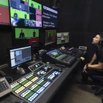 A person monitors multiple screens and a control panel in a studio, preparing for a live broadcast on green screen content.