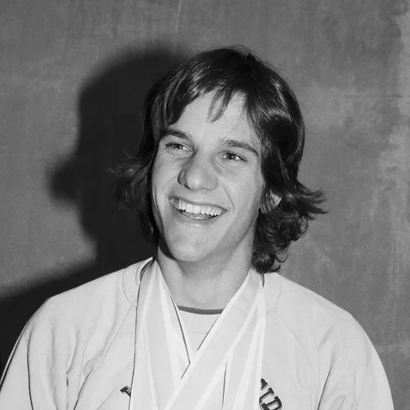 Eric Heiden grins broadly in a black-and-white portrait, medals draped around his neck.