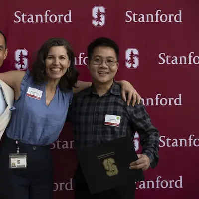 Three individuals celebrate with arms around each other, holding certificates in front of a Stanford University backdrop.