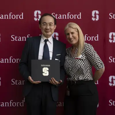 Two individuals pose for a photo holding a certificate in front of a Stanford-themed backdrop during an event.