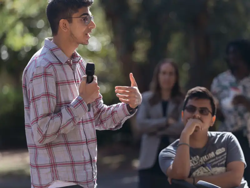A student speaks into a microphone at an outdoor campus event while others listen attentively in the background.
