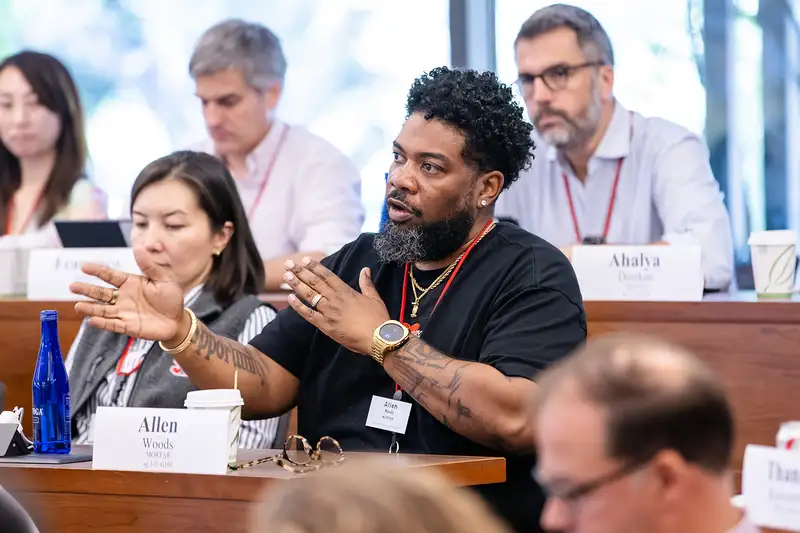 A participant actively engaging in discussion during a meeting, seated among others in a conference room with name tags visible.
