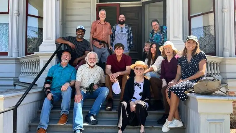 A group of 12 people sits on the steps of a house, smiling together in a casual outdoor gathering.